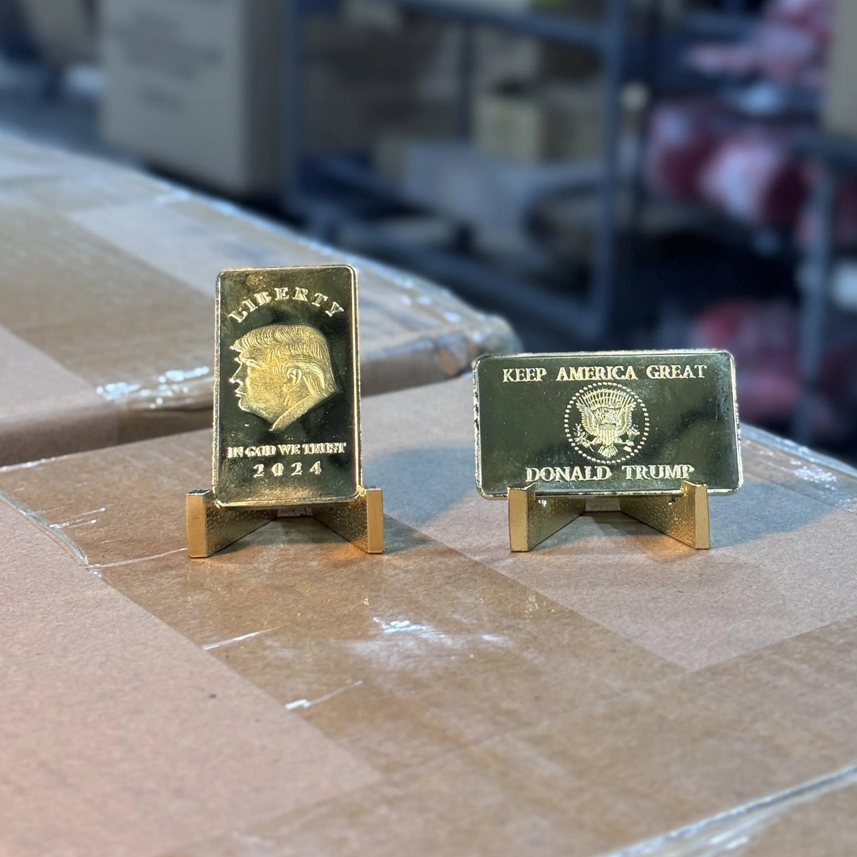 Trump Liberty 2024 gold-plated bars on stands on a packing table, front and reverse sides shown