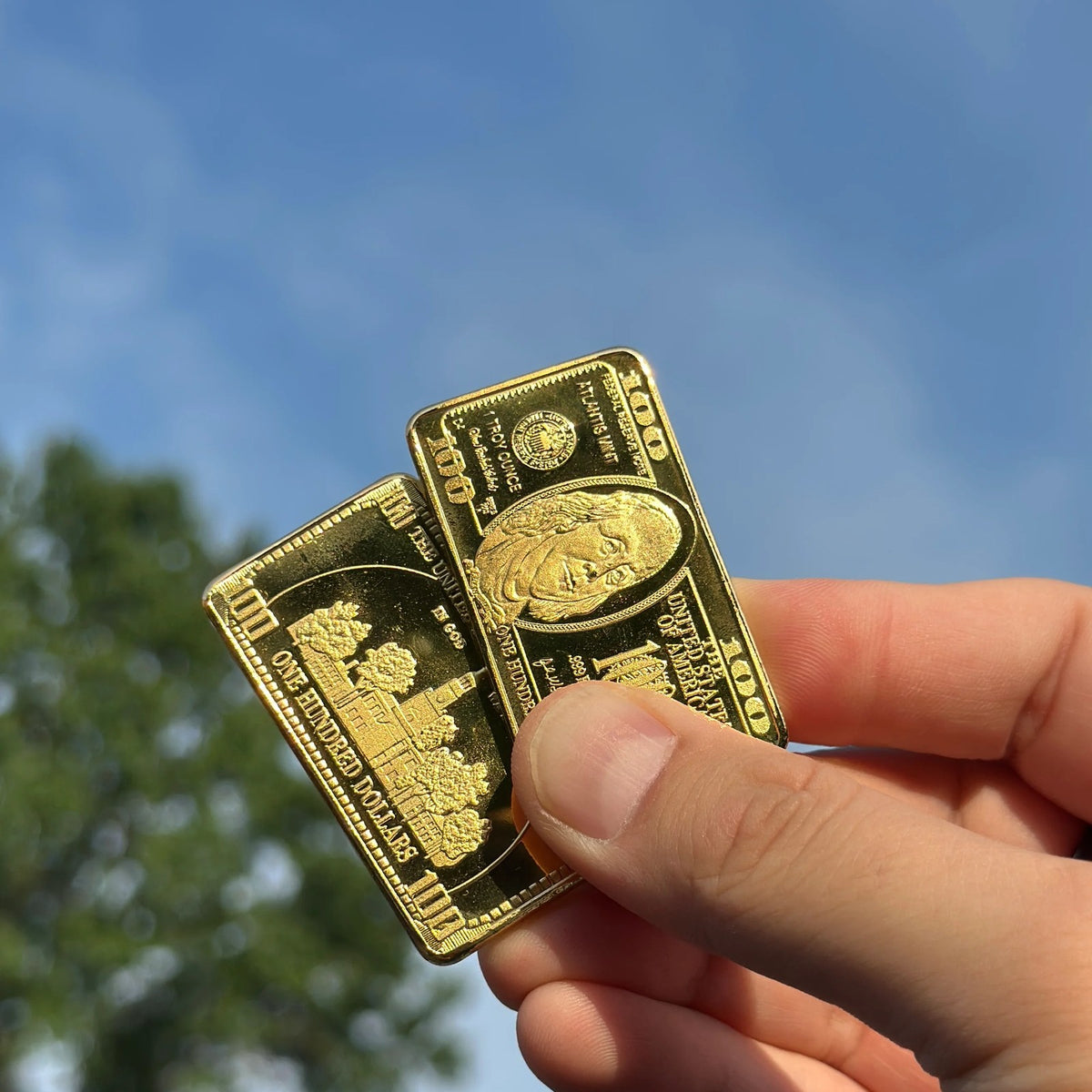 Two Ben Franklin $100 gold-plated bars held between fingers against the sky for size