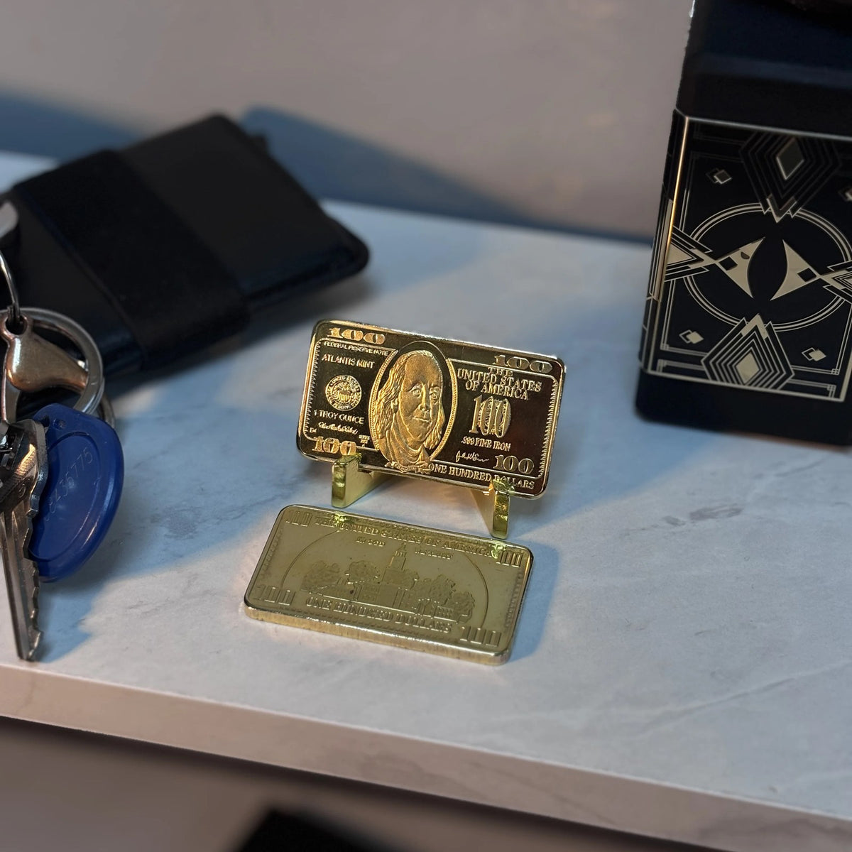 Ben Franklin $100 gold-plated bar on a desk stand next to keys and a wallet
