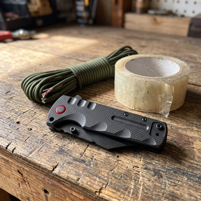 Black Vantage Knife folded on a garage workbench beside paracord and packing tape