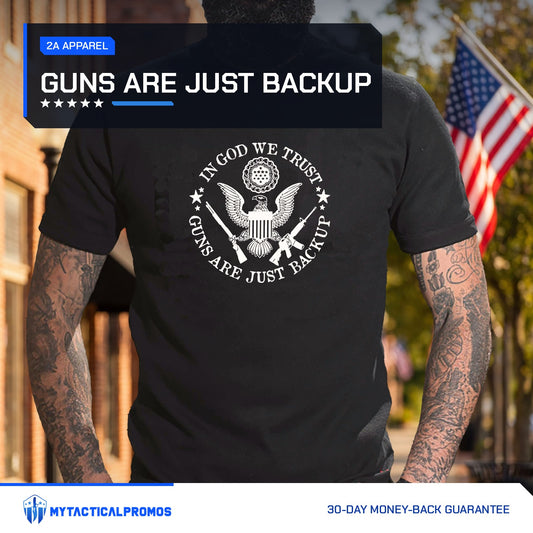 Guns Are Just Backup T-Shirt worn by a man in front of a small-town main street lined with flags