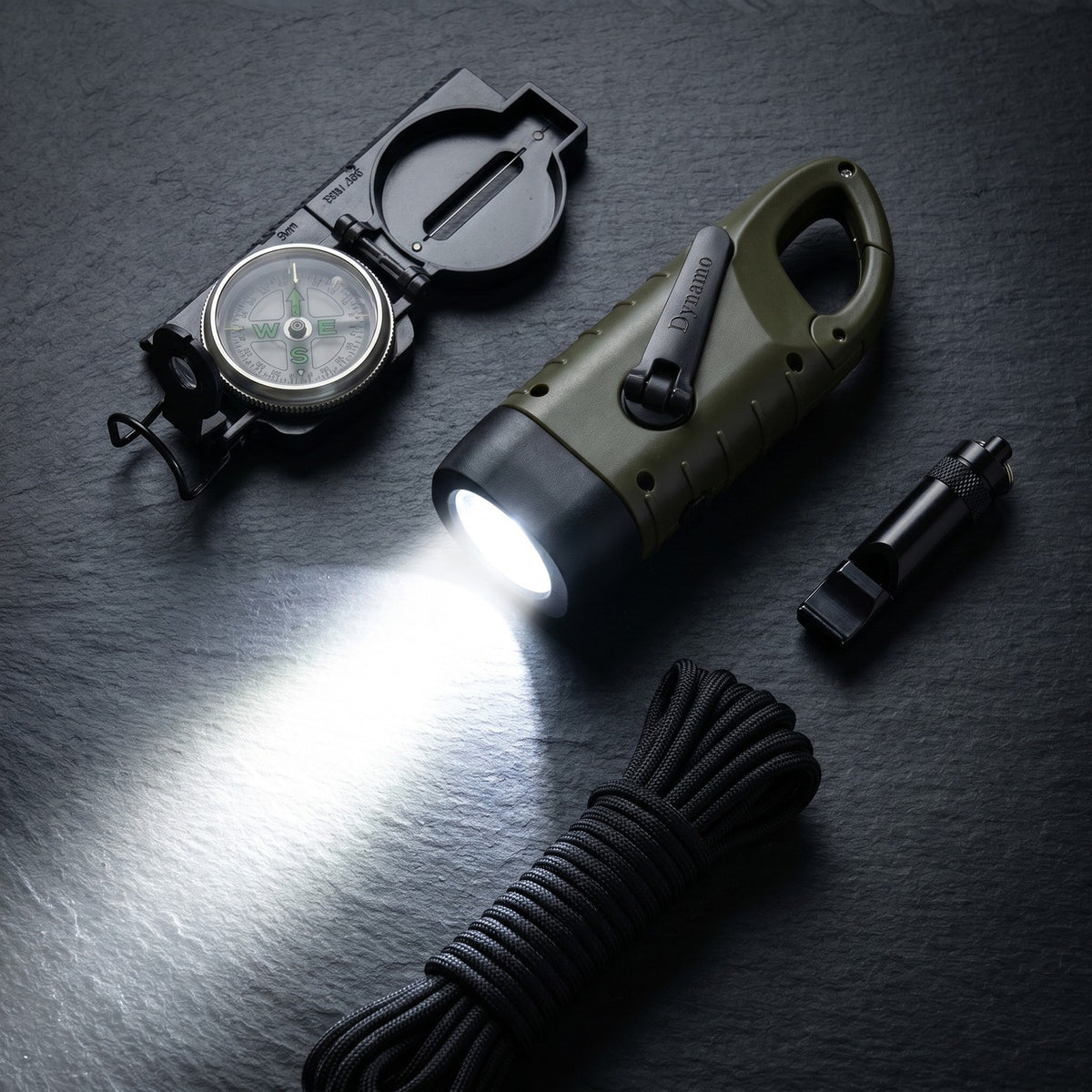Hand Crank Flashlight lit on dark slate beside a compass, whistle and paracord