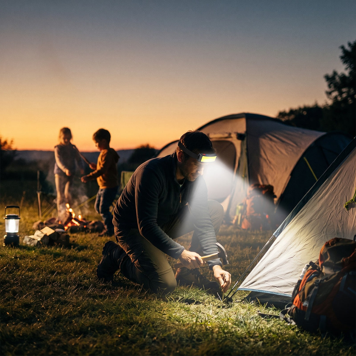 Man wearing the Headlamp while pitching a tent at dusk, kids by the campfire