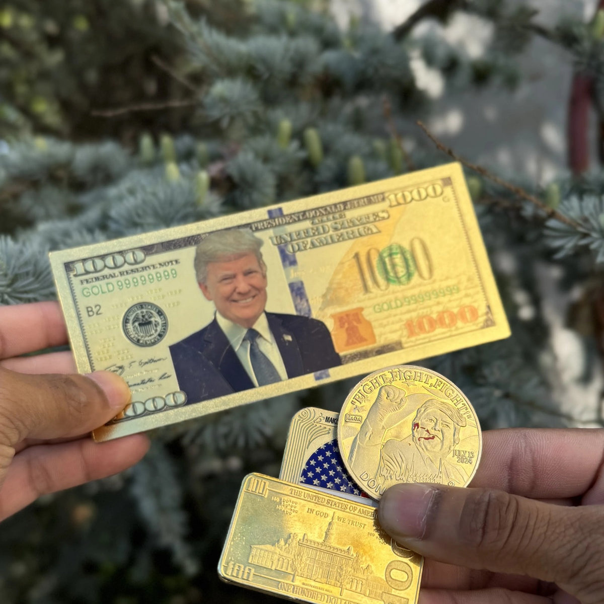 Trump $1000 gold foil novelty bill held outdoors by an evergreen, other keepsakes sold separately