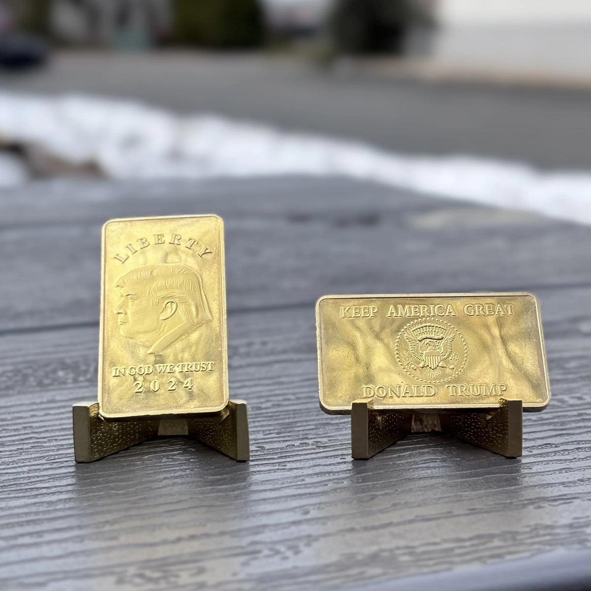 Trump Liberty 2024 gold-plated bars on stands on an outdoor table in winter, front and reverse shown