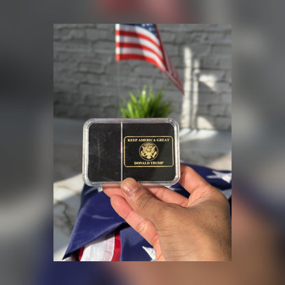 Trump Liberty 2024 black and gold bar reverse held in front of a folded American flag