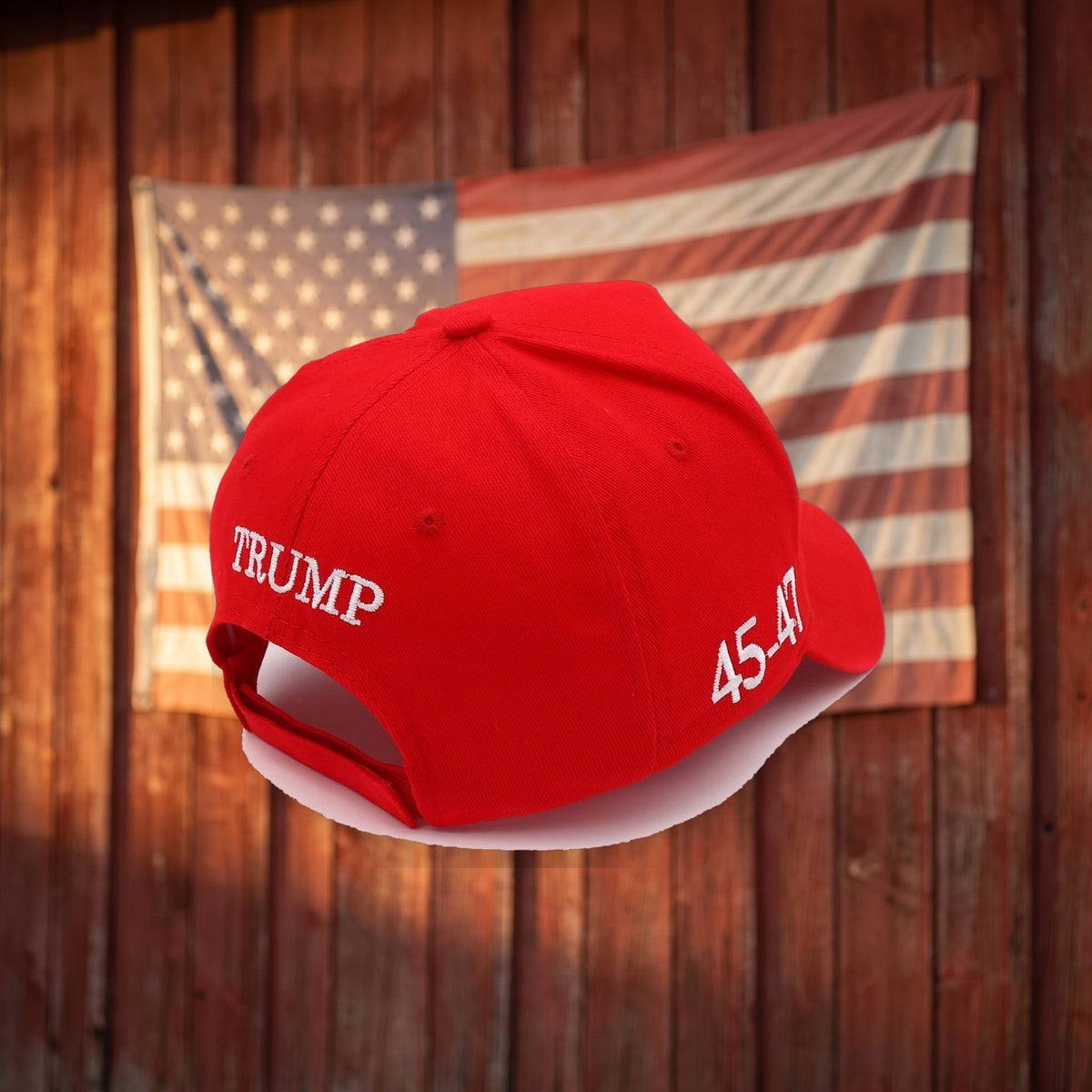 Red Trump USA Hat back view in front of a flag on a barn wall
