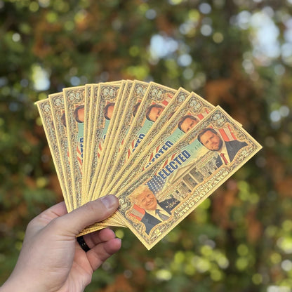 Several Trump Vance $2 gold foil novelty bills fanned out in one hand outdoors