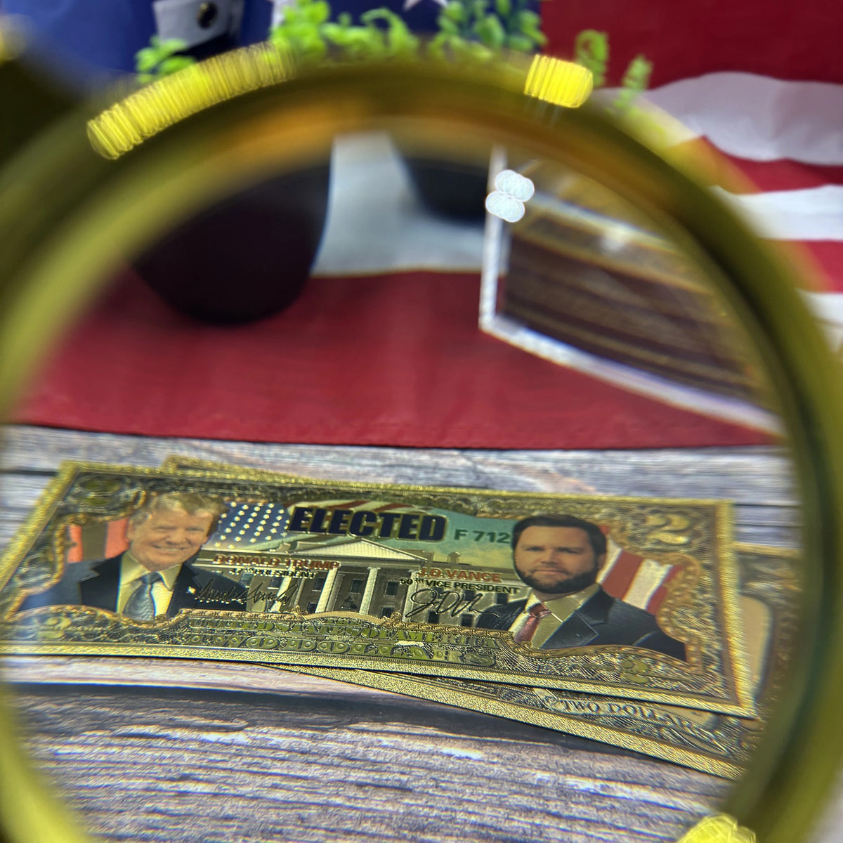 Trump Vance $2 gold foil novelty bill seen through a magnifying glass in front of a flag