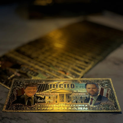 Trump Vance $2 gold foil novelty bill on a dark desk