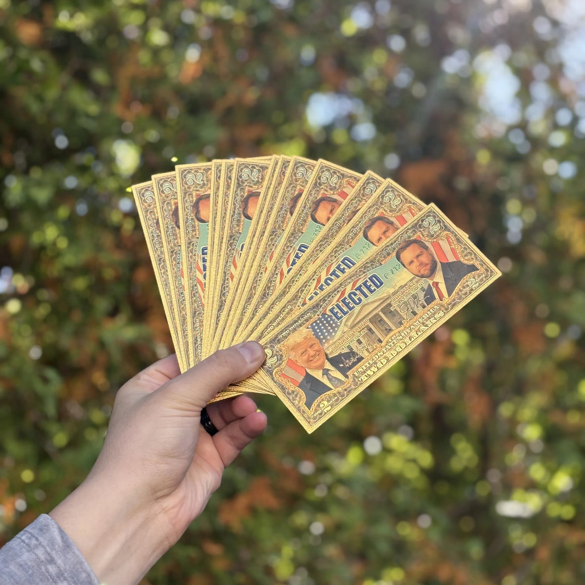 Several Trump Vance $2 gold foil novelty bills fanned out against autumn trees